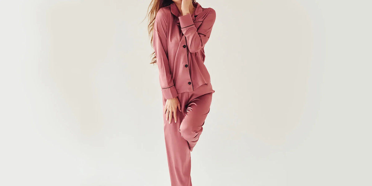 Banner für die Kategorie „Damen Nachtwäsche“ im Boxxers Online Shop. Eine Frau trägt einen zweiteiligen, roséfarbenen Pyjama mit langen Ärmeln und langer Hose und posiert vor einem hellen Hintergrund.