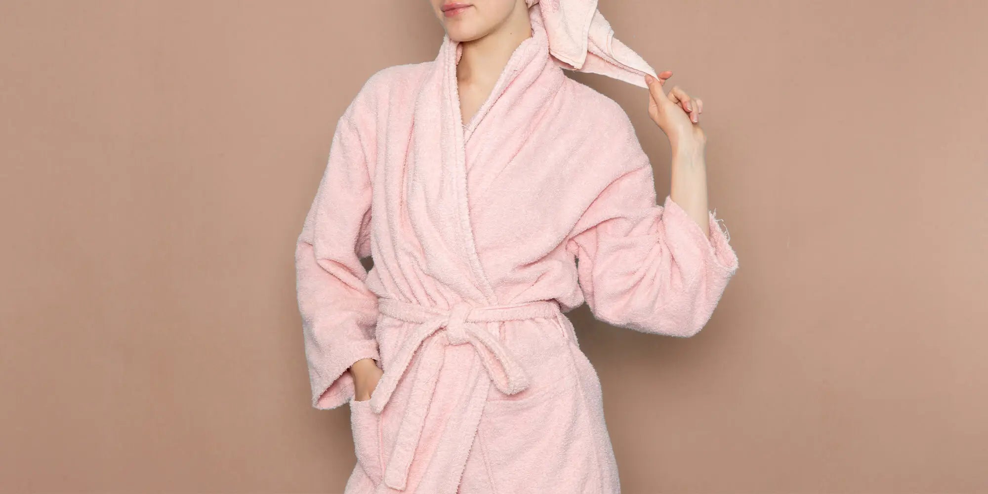 Banner für die Kategorie „Damen Bademäntel“ im Boxxers Online Shop. Eine Frau trägt einen rosa Frottee-Bademantel und hat ein Handtuch um den Kopf gewickelt, während sie vor einem beigebraunen Hintergrund steht.