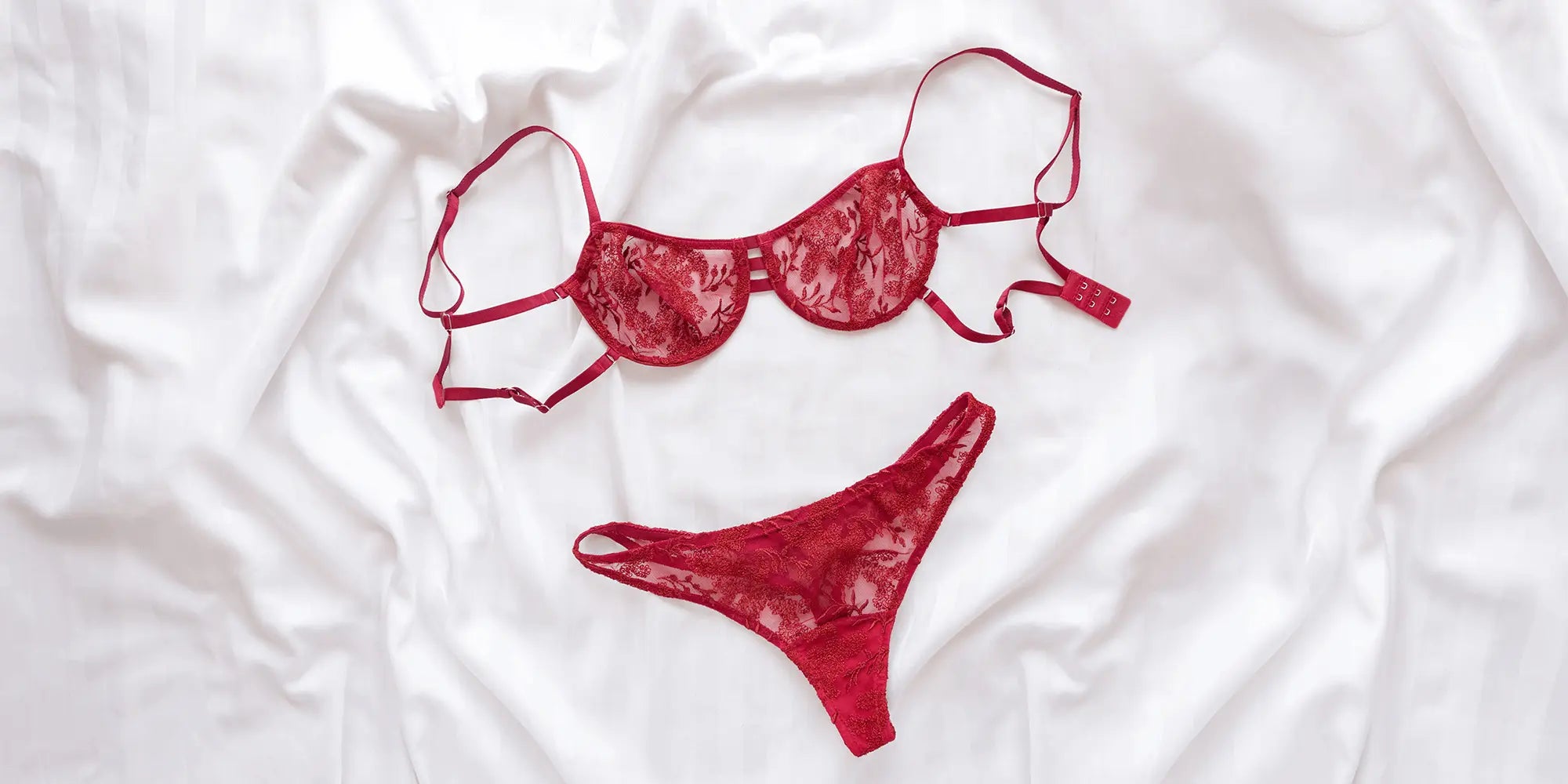 Banner für die Kategorie „Damen Unterwäsche Sets“ im Boxxers Online Shop. Ein rotes Spitzen-Unterwäsche-Set aus BH und Slip liegt dekorativ auf weißer Bettwäsche.