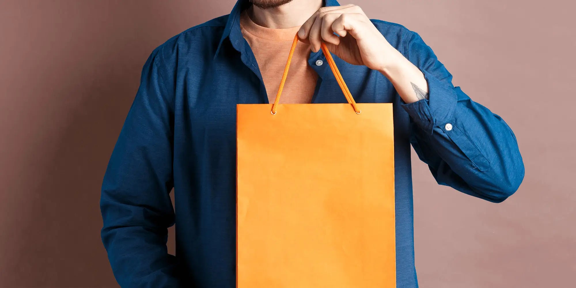 Banner für die Kategorie „SALE für Herren“ im Boxxers Online Shop. Ein Mann trägt ein blaues Hemd über einem orangefarbenen T-Shirt und hält eine große, orange Einkaufstüte vor einem braunrosafarbenen Hintergrund.
