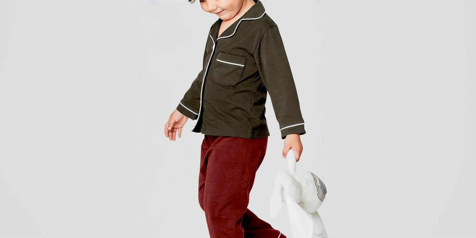 Banner für die Kategorie „Kinder Pyjamas“ im Boxxers Online Shop. Ein Kind trägt einen zweiteiligen Pyjama mit dunkelgrünem Oberteil und bordeauxroter Hose und hält ein weißes Kuscheltier, während es vor einem hellen Hintergrund läuft.