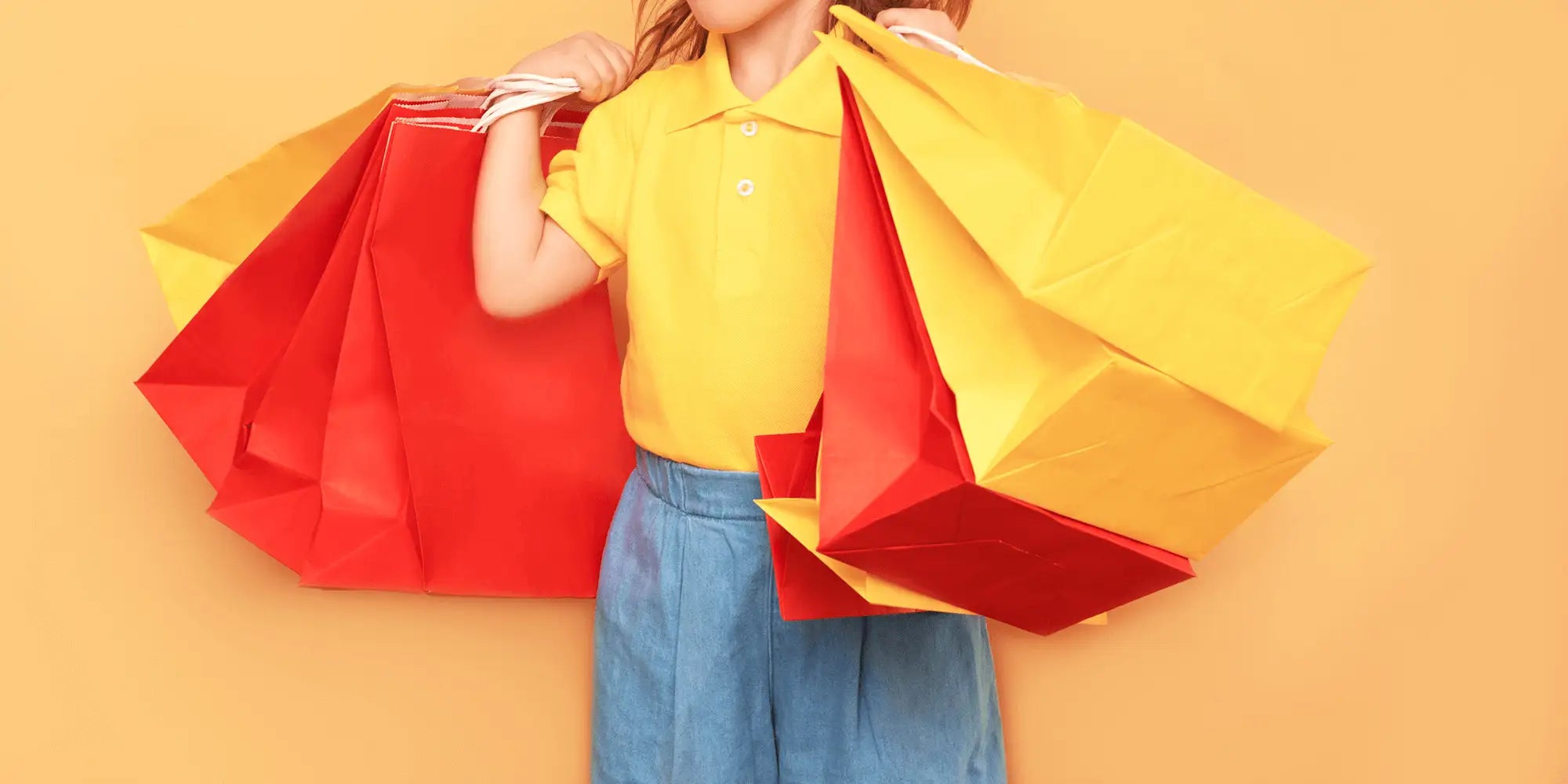 Banner für die Kategorie „SALE für Kinder“ im Boxxers Online Shop. Ein Kind trägt ein gelbes Poloshirt und blaue Hose und hält mehrere große, rote und gelbe Einkaufstüten vor einem einfarbigen, orangefarbenen Hintergrund.